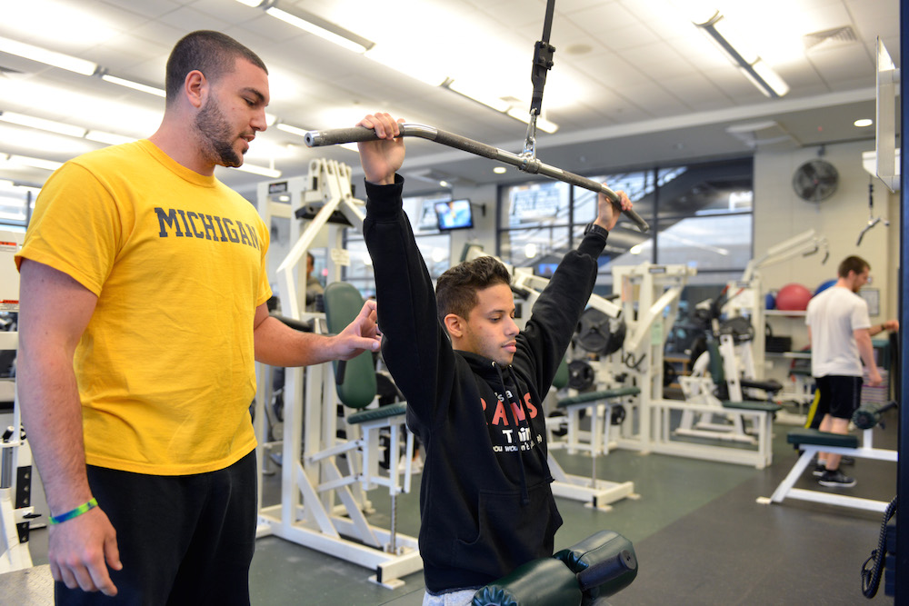 A male student uses exercise equipment while a personal trainer looks on